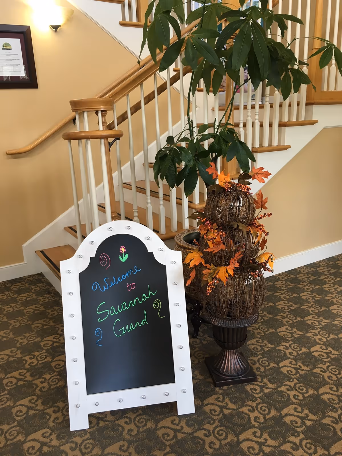 Entrance lobby area with a decorative welcome chalkboard sign, potted plants, and a wooden staircase.