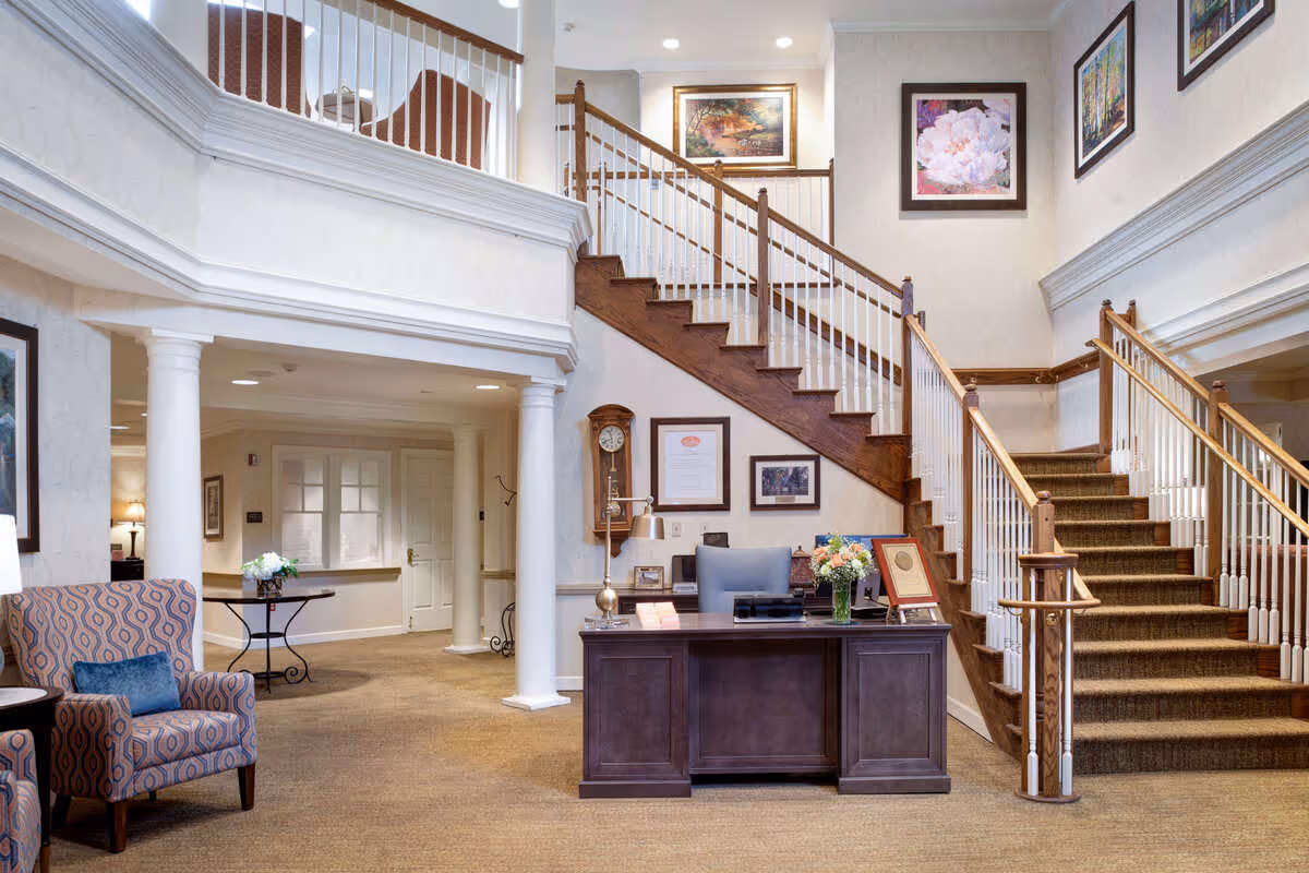A spacious and well-lit interior area of a senior living facility featuring a wooden reception desk with a chair, a vase of flowers, and framed certificates on the wall behind it. To the right, there is a carpeted staircase with wooden handrails leading to an upper floor. The walls are decorated with framed artwork. On the left side, there is a patterned armchair with a blue cushion and a small table with a flower arrangement. The area has white columns and beige carpeting.