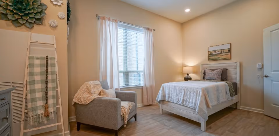 Bright furnished bedroom with a single bed, armchair, window with sheer curtains, and a decorative blanket ladder.