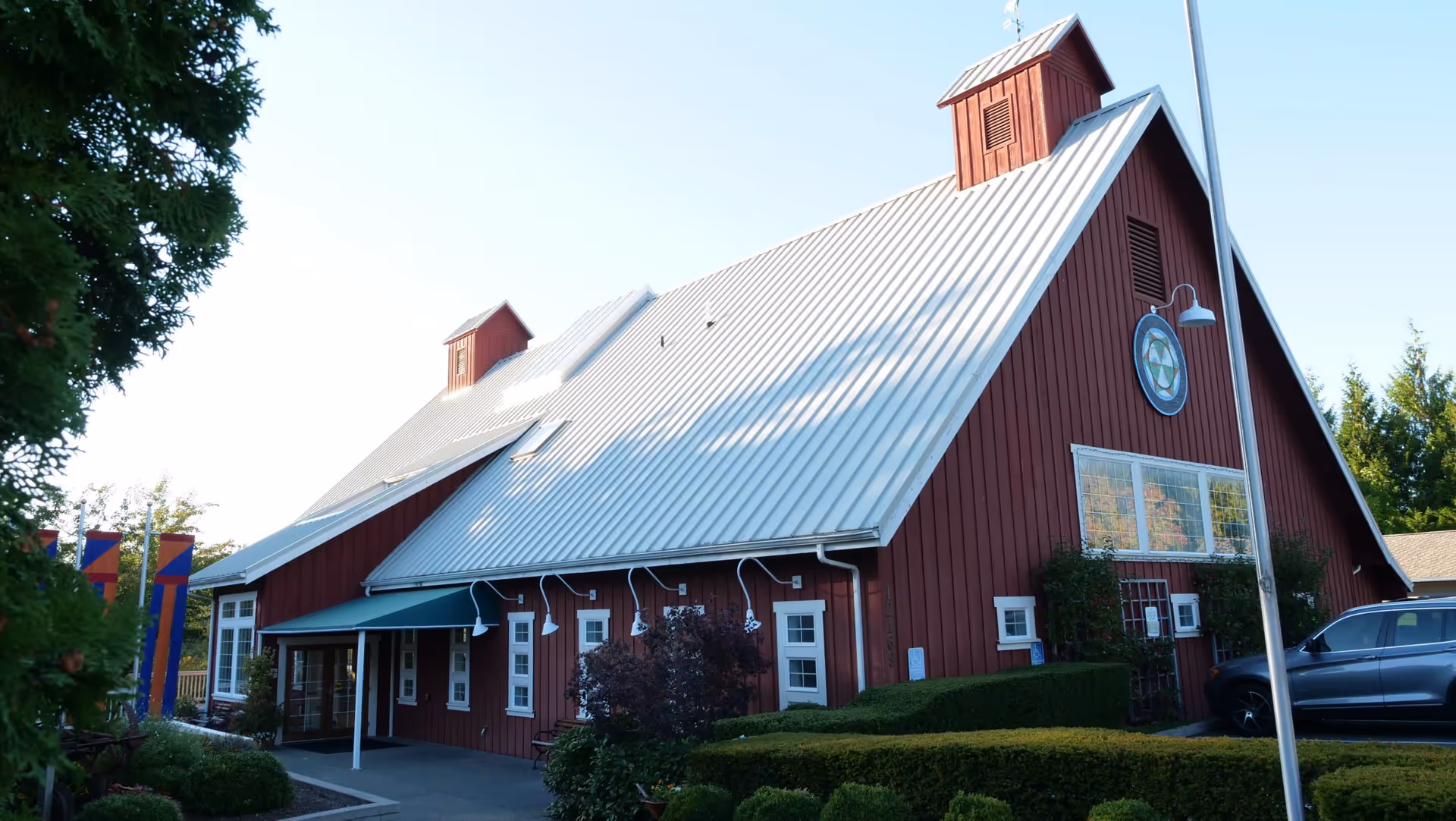 Exterior view of a large red barn-style building with a white metal roof, surrounded by greenery and bushes. There is a car parked on the right side and a flagpole in front of the building. The building has multiple small windows and a circular decorative element on the front wall.
