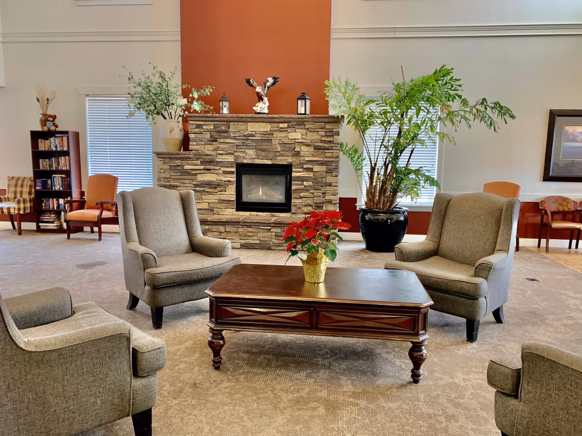 A cozy living room area with four gray upholstered armchairs arranged around a wooden coffee table with a red poinsettia plant on it. Behind the seating area is a stone fireplace with decorative items on the mantel, flanked by two windows with blinds. There is a large potted plant to the right and a bookshelf with books and a teddy bear to the left. Additional chairs and framed artwork are visible in the background.