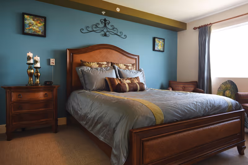 A cozy bedroom with a wooden bed frame and headboard, neatly made with gray and gold bedding and multiple decorative pillows. To the left of the bed is a wooden nightstand with three drawers, holding two lit candles on brass candle holders and a small clock. The wall behind the bed is painted teal and decorated with two abstract paintings and a decorative wrought iron piece. To the right, a window with gray curtains lets in natural light, and there is a cushioned chair and a decorative fan-shaped object near the window.