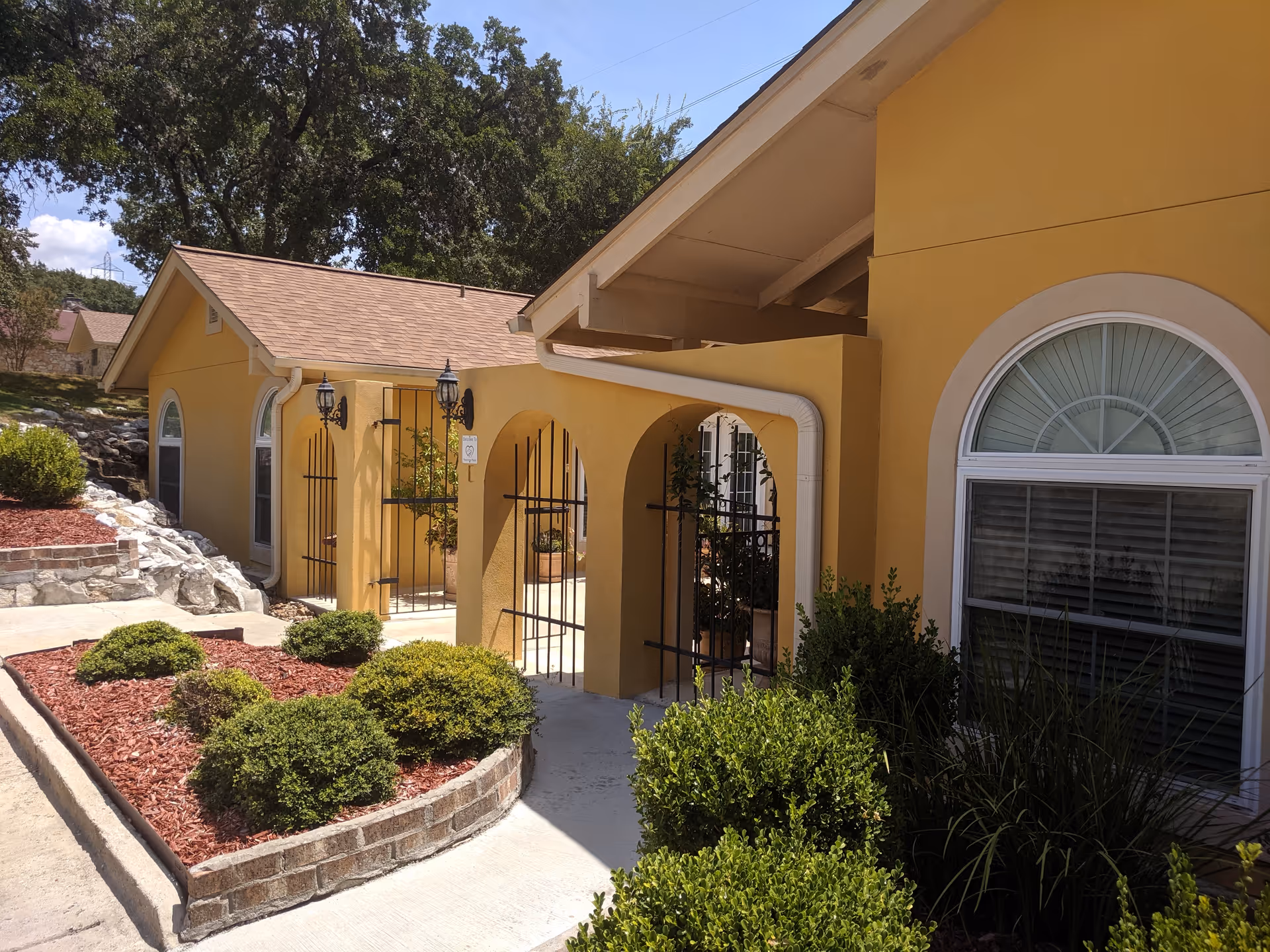 Exterior view of a yellow building with arched entryways and black metal gates. The building has a tan roof and arched windows with white trim. There are neatly trimmed bushes and landscaped areas with red mulch along a concrete walkway. Trees and a clear blue sky are visible in the background.