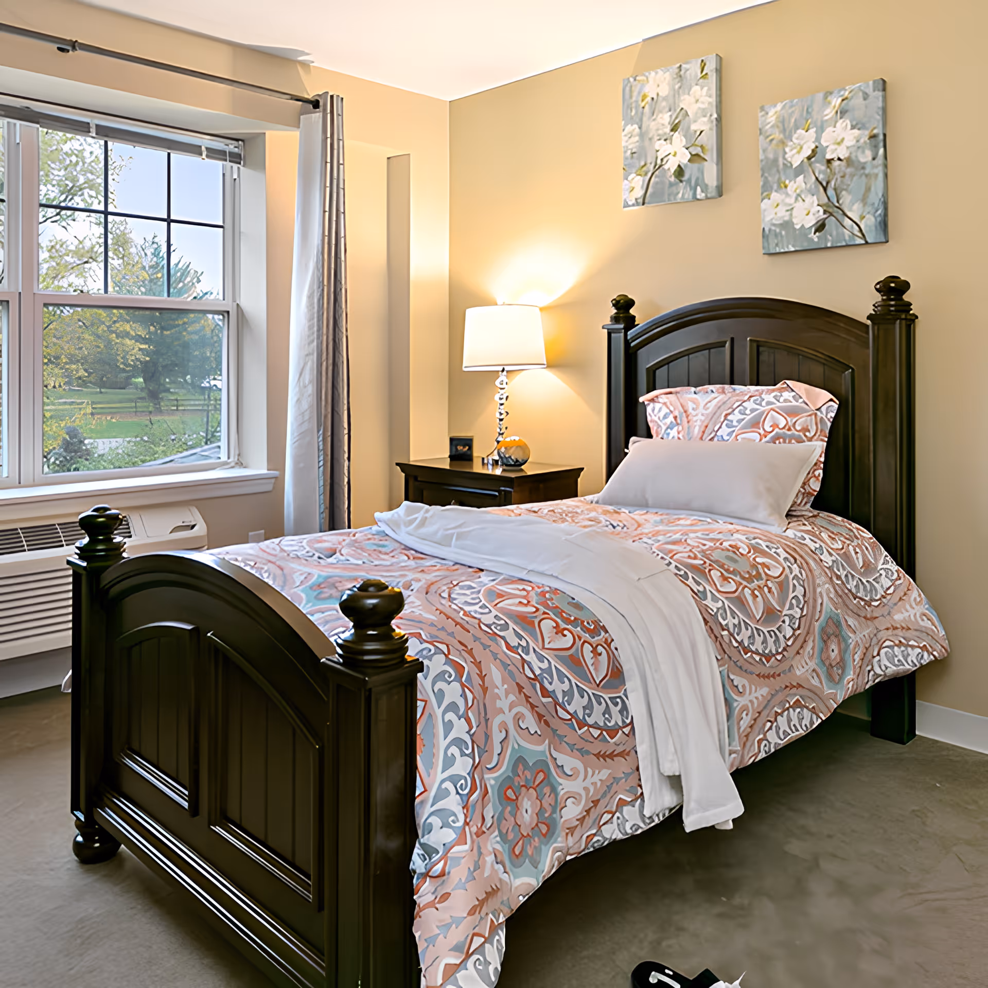 A neatly made single bed with a dark wooden frame and colorful patterned bedding in a bedroom. There is a nightstand with a lamp and a small decorative item next to the bed. Two floral paintings hang on the beige wall above the bed. A large window with curtains lets in natural light and shows greenery outside.
