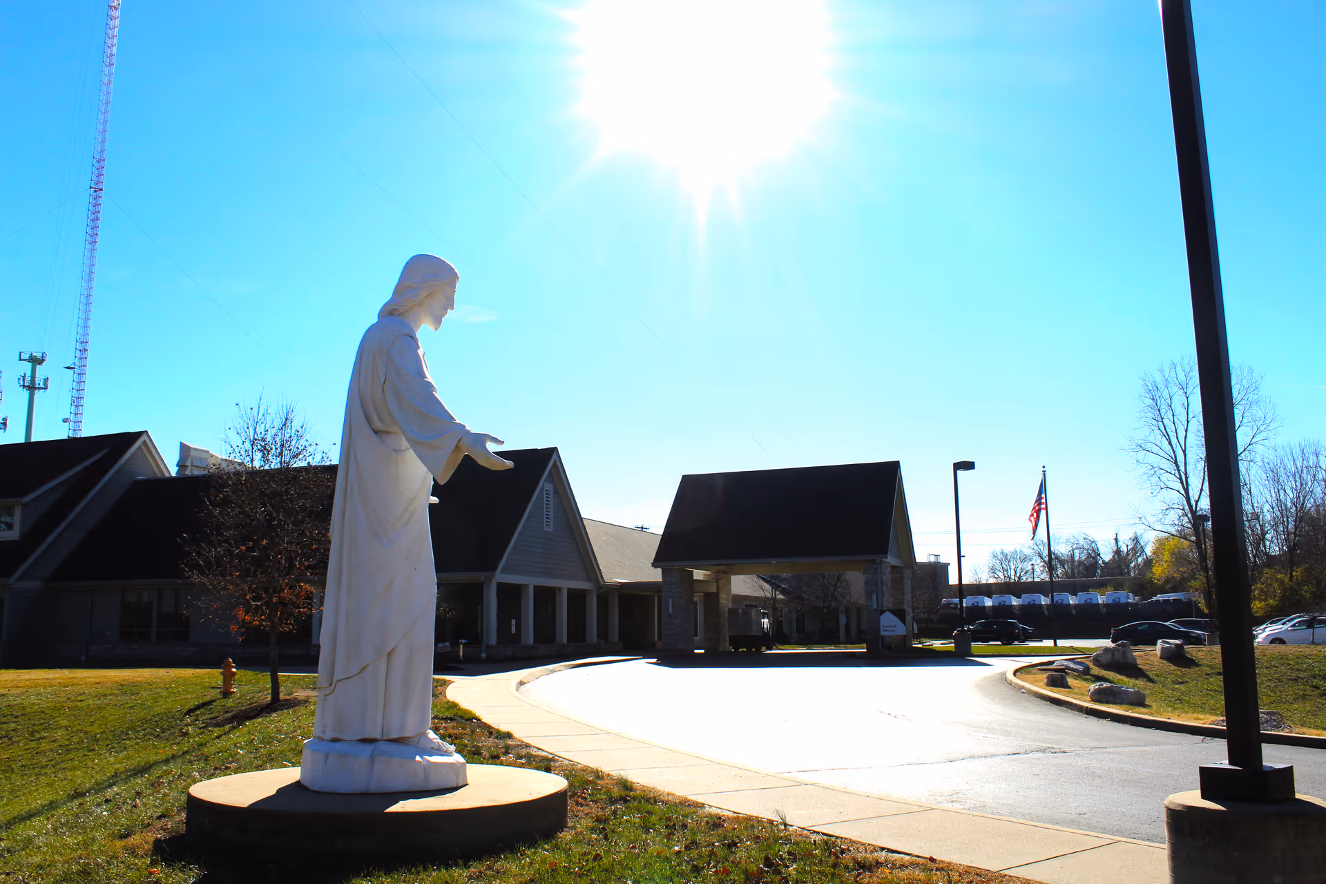 A white religious statue on a pedestal stands on the lawn beside the driveway leading to the front entrance of a senior living building under a bright sun.