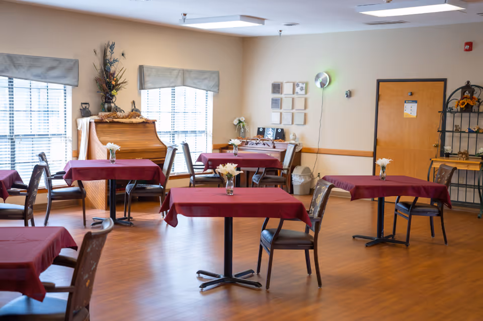 A dining room with several tables draped in burgundy tablecloths, chairs, a piano by the windows, and small vases of flowers on each table.