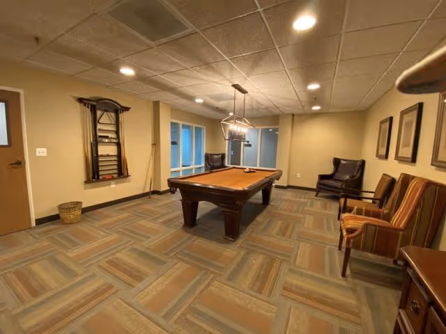 A recreational room with a pool table in the center, two chairs against the right wall, a wall-mounted rack holding pool cues on the left wall, and a modern light fixture hanging above the pool table. The room has a patterned carpet floor, beige walls, and a drop ceiling with recessed lighting.