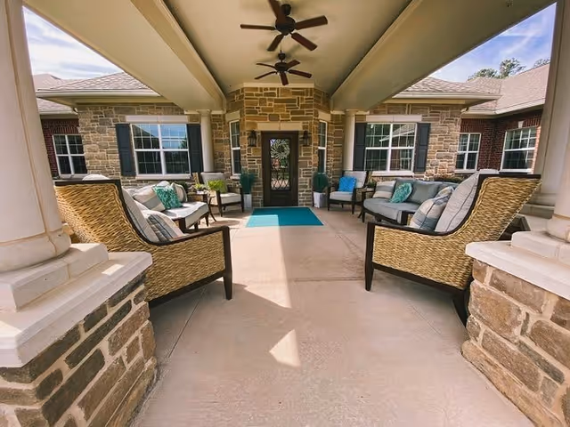Covered outdoor seating area with wicker chairs and cushioned sofas arranged around a central walkway leading to a door. The area has stone pillars and walls, ceiling fans, and windows with black shutters.