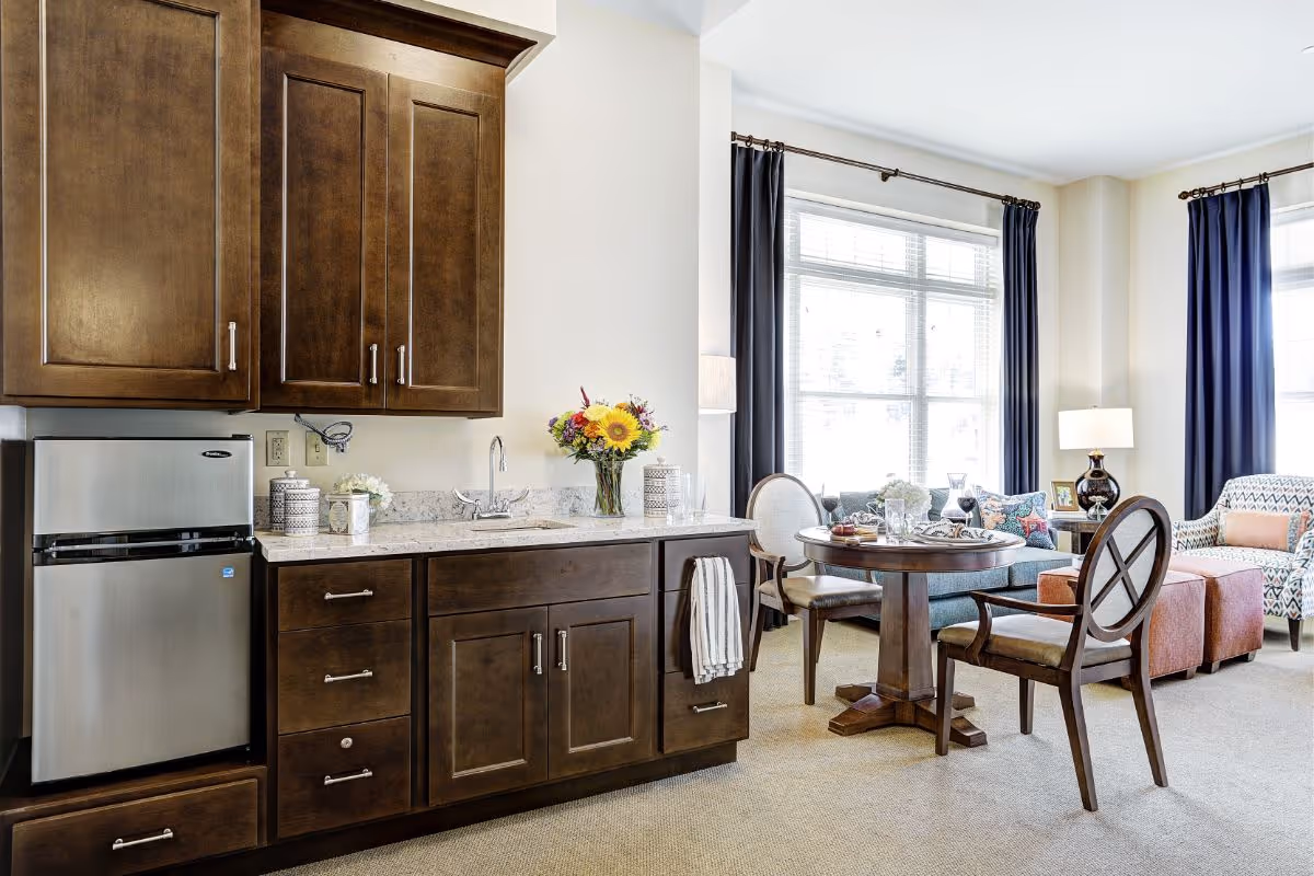 Well-lit senior living suite with a small kitchenette (dark cabinets and mini-fridge) opening to a dining table and comfortable seating by large windows.