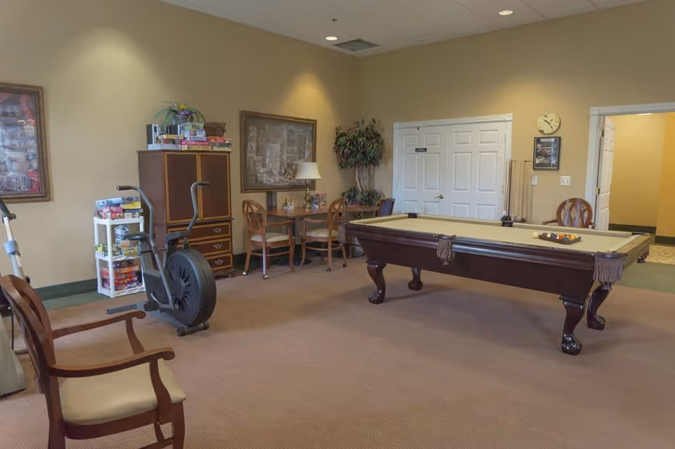 A recreational room with a pool table in the center, a stationary exercise bike, a small table with chairs, a cabinet with board games stacked on top, and framed artwork on the walls. The room has beige walls and carpeted floor, with a clock and a plant near a set of double doors.