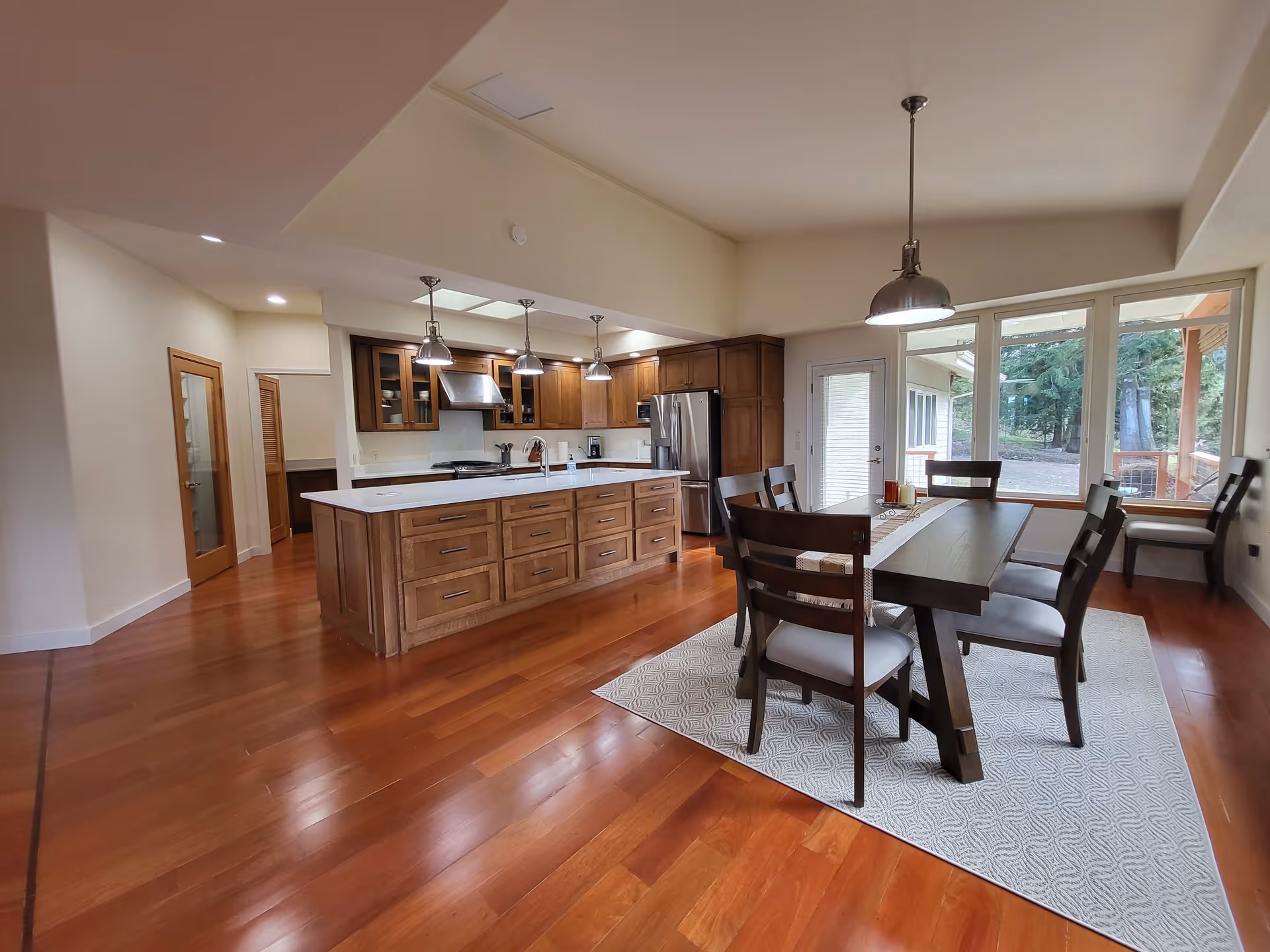 Spacious kitchen and dining area with wooden floors and cabinetry. The kitchen features a large island with multiple drawers, stainless steel appliances, and three pendant lights hanging above the island. The dining area has a dark wooden table with six chairs placed on a patterned rug, next to large windows and a glass door that provide a view of outdoor greenery.