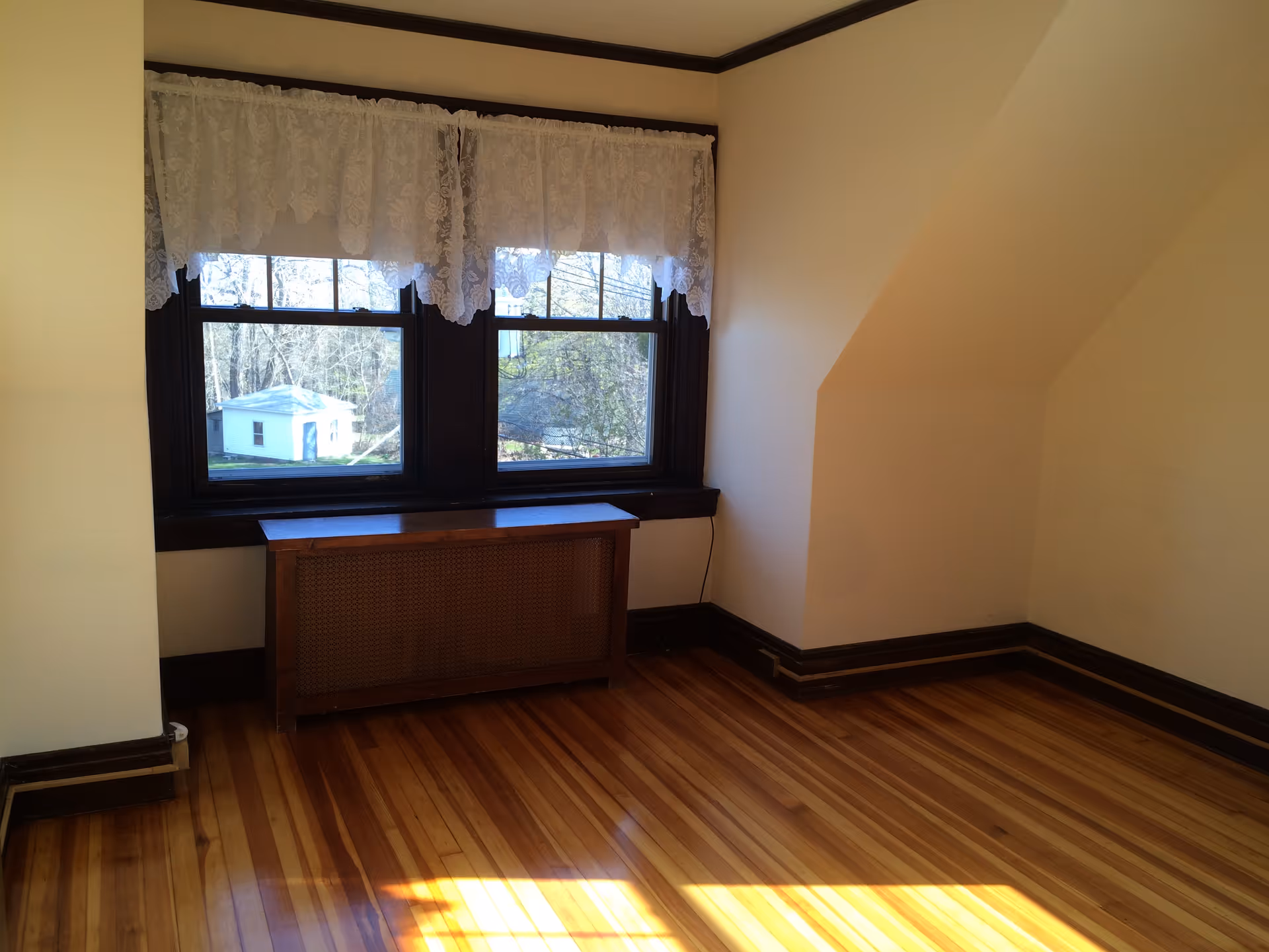 Empty sunlit room with hardwood floors, lace-curtained double windows, and a wooden radiator cover.