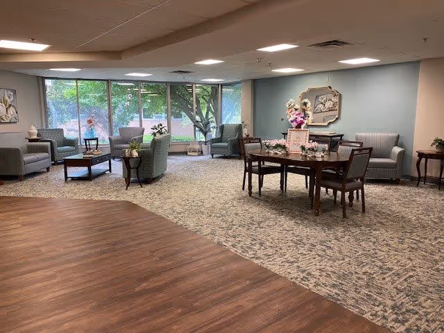 A spacious and well-lit common area in a senior living facility with large windows showing greenery outside. The room features multiple seating arrangements including armchairs, a sofa, and a wooden table with chairs. The floor is a combination of wood and carpet, and the walls are decorated with framed artwork and floral arrangements.