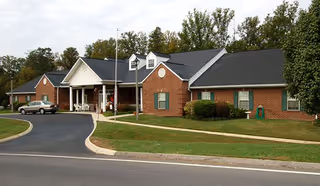 Exterior view of a single-story brick building with a covered entrance, surrounded by a well-maintained lawn and trees in the background. A car is parked near the entrance on a paved driveway.
