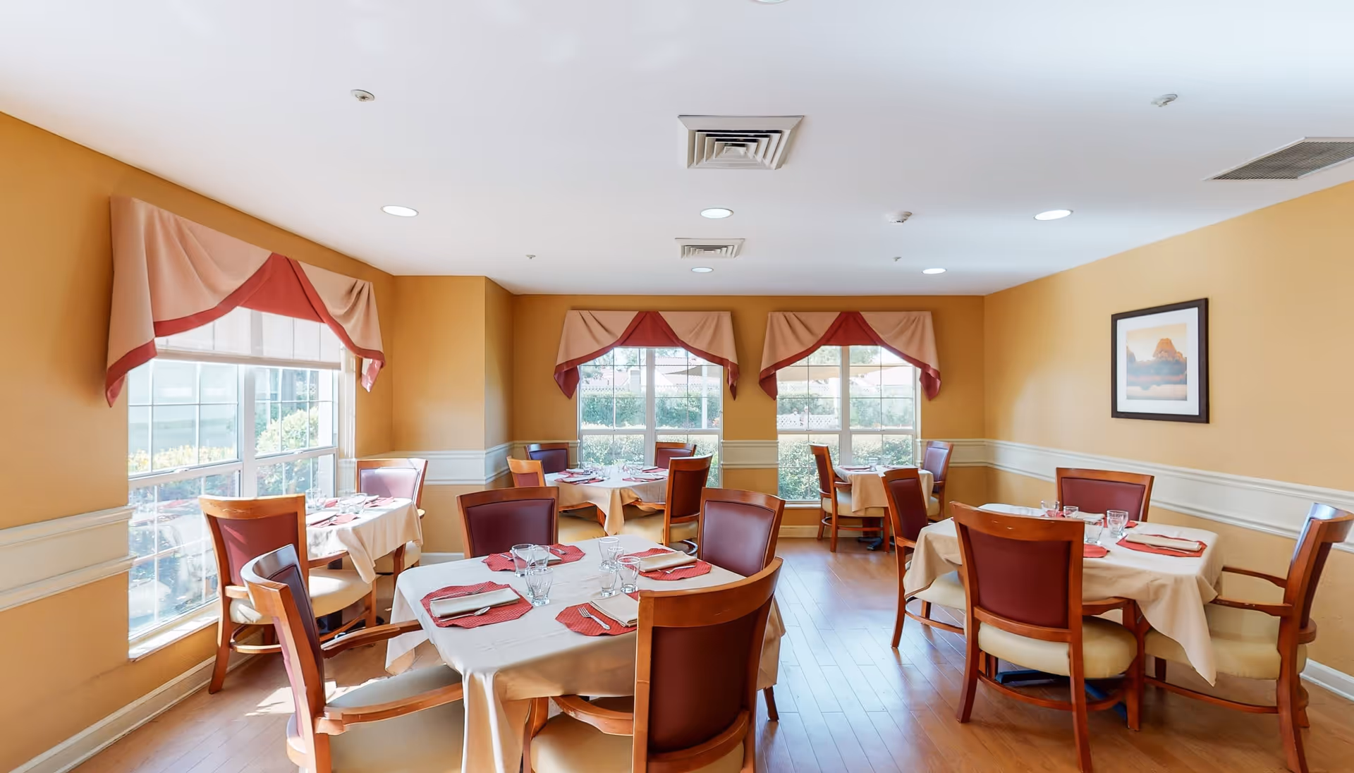 A bright dining room with several round and square tables covered with beige tablecloths and red napkins. Each table is set with glasses and silverware. The room has large windows with red and beige valances, wooden chairs with red cushions, light brown walls, and hardwood floors. A framed picture hangs on one wall.