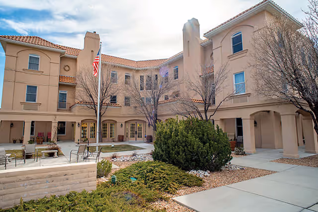 Exterior view of a multi-story senior living facility building with beige stucco walls and a red tile roof. The courtyard features leafless trees, green shrubs, a flagpole with an American flag, and outdoor seating areas with chairs and tables. The sky is partly cloudy.