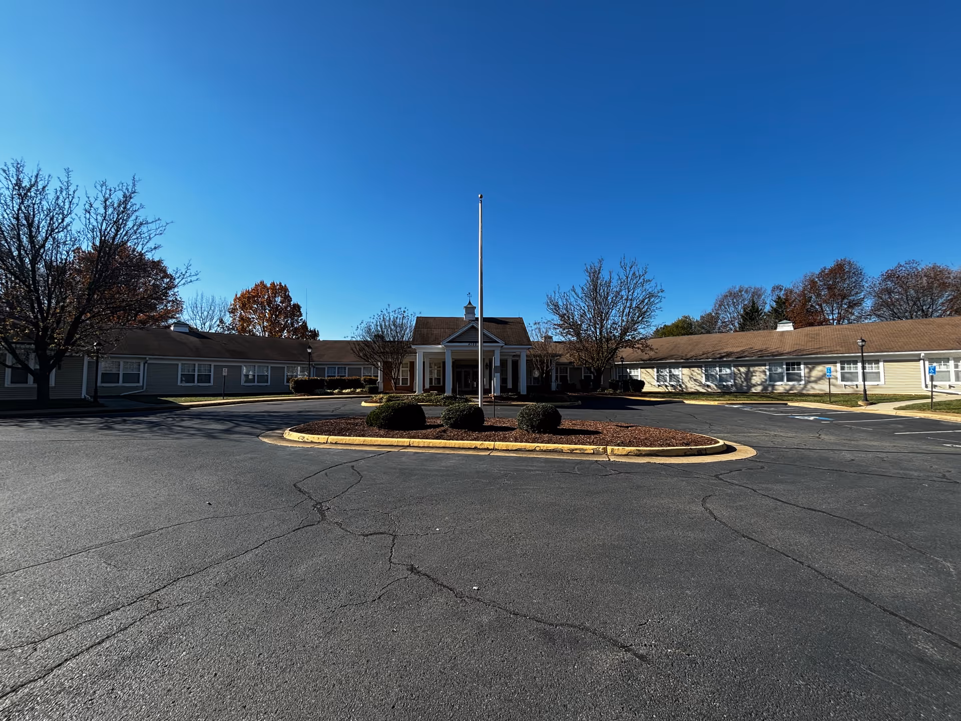 Exterior view of a single-story senior living facility building with a central entrance featuring white columns and a small cupola on the roof. The building is surrounded by a paved circular driveway with a landscaped island in the center containing bushes and a flagpole. Trees with autumn foliage are visible around the building under a clear blue sky.