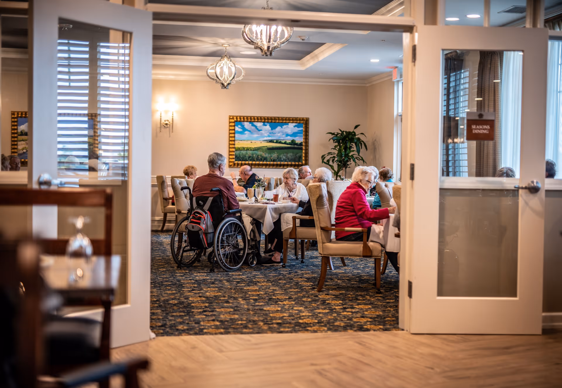View through open double doors into a dining room where elderly residents are seated at tables enjoying a meal. The room is warmly lit with chandeliers and wall sconces, decorated with a large framed landscape painting and a potted plant. One resident is in a wheelchair.