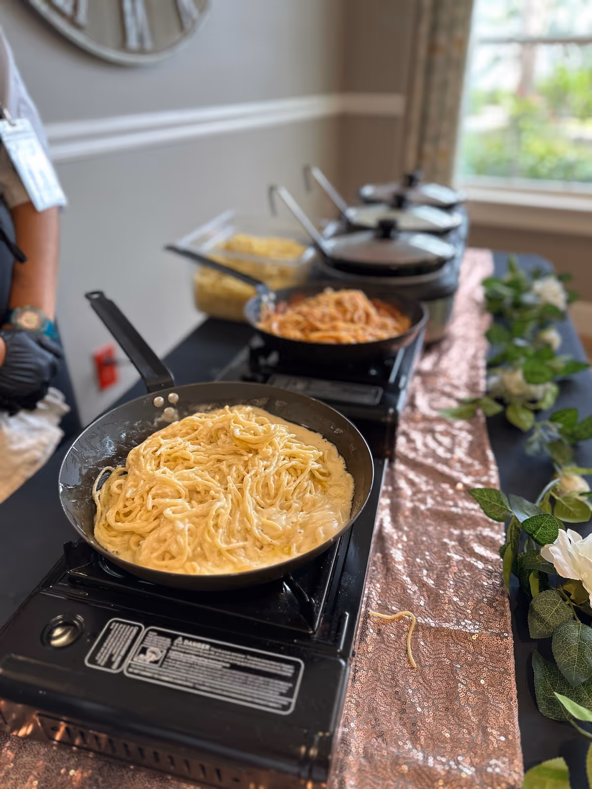 Skillet of creamy pasta on a buffet table with additional pans of pasta and sauces on portable burners and floral decorations.