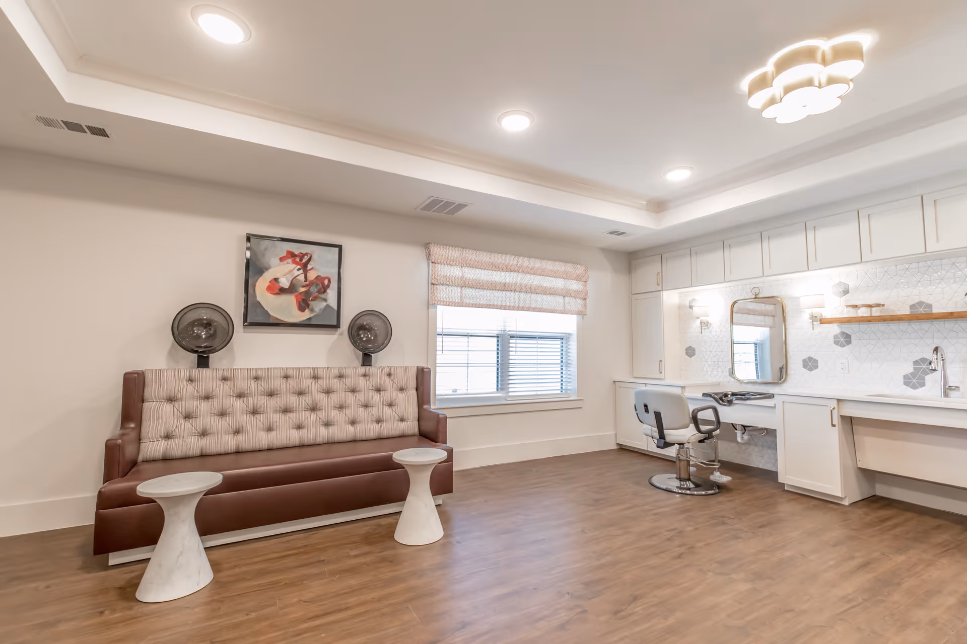 Bright salon-style interior with a tufted bench, two small white tables, and a styling chair and mirror at a built-in counter with cabinets.