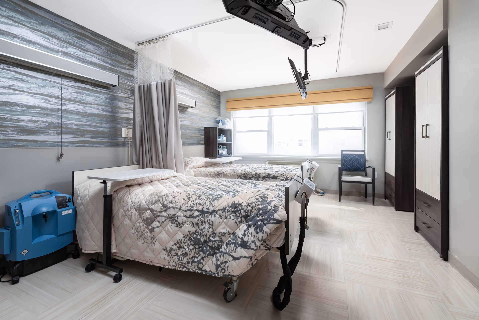 A bright and clean room with two hospital-style beds separated by a privacy curtain. Each bed has a patterned quilt and an overbed table. There is a blue medical device on the left side of the room, a chair near the window, and two wardrobes on the right side. A ceiling-mounted TV is positioned above the beds. The room has large windows with a beige valance allowing natural light to fill the space.