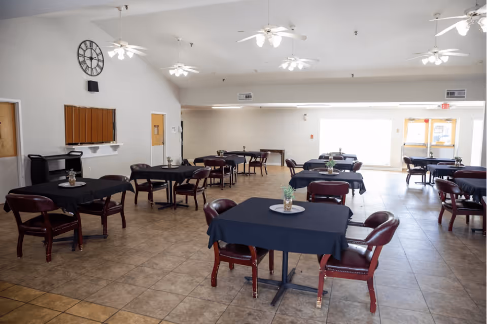 A spacious dining room with multiple tables covered in black tablecloths, each surrounded by four maroon chairs. Small decorative plants are placed in the center of each table. The room has tiled floors, white walls, ceiling fans with lights, and a large clock on one wall. There are doors and windows letting in natural light at the far end of the room.