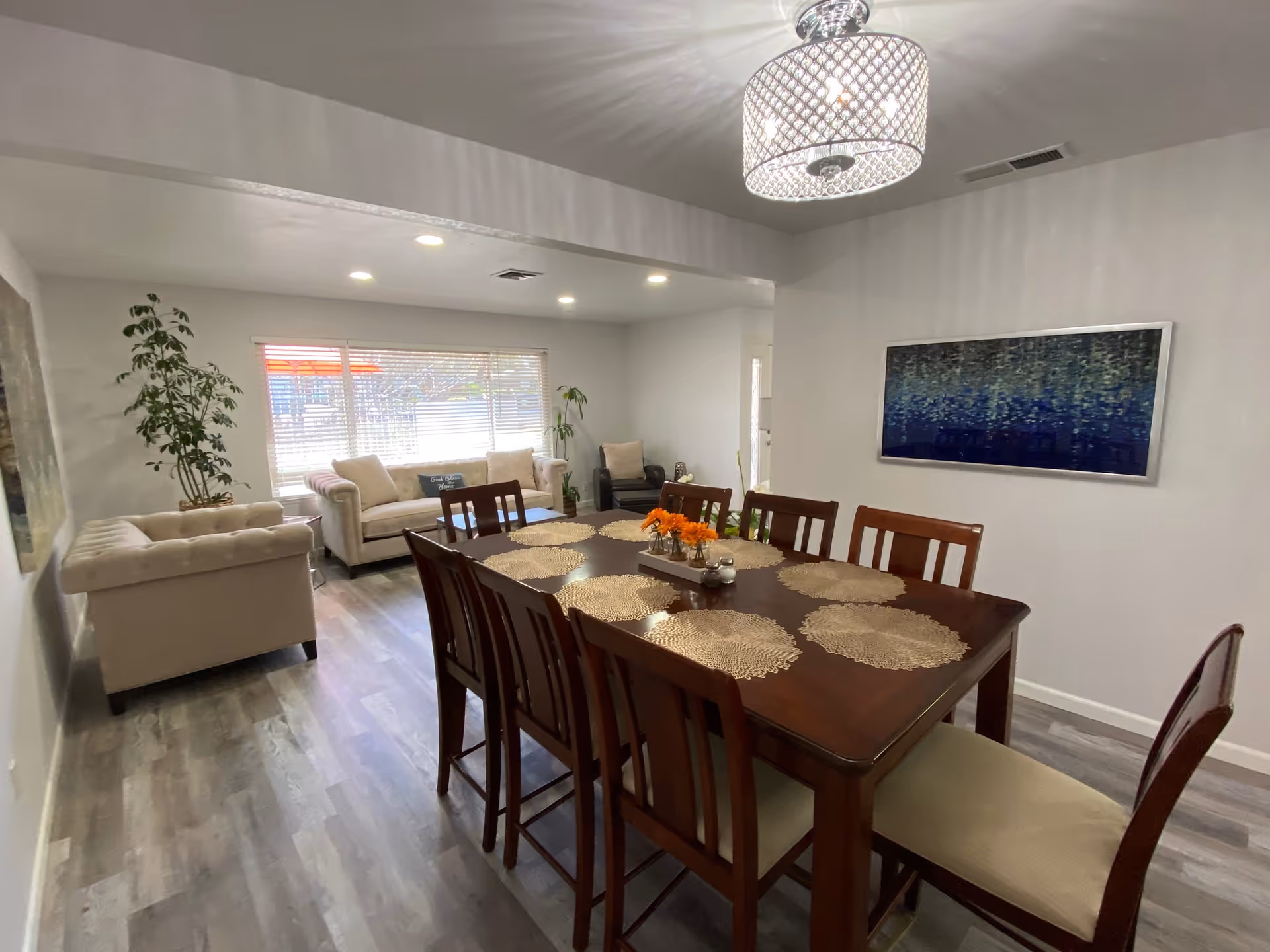 A bright and spacious living and dining area with a wooden dining table set for eight, beige cushioned chairs, two beige sofas, a single armchair, potted plants, a large window with blinds, a modern ceiling light fixture, and a blue abstract painting on the wall.