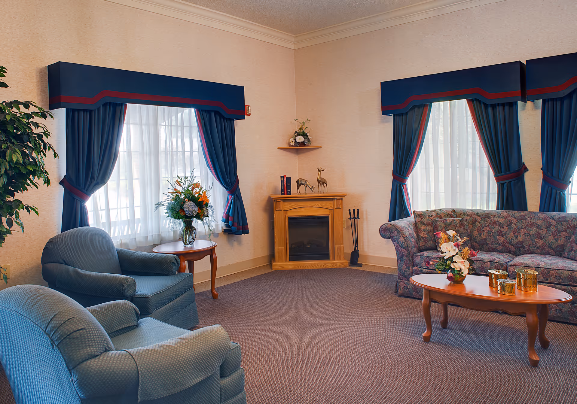 Cozy common living room with upholstered armchairs and a floral-upholstered sofa, a coffee table with flowers, a small fireplace, and blue draped windows.