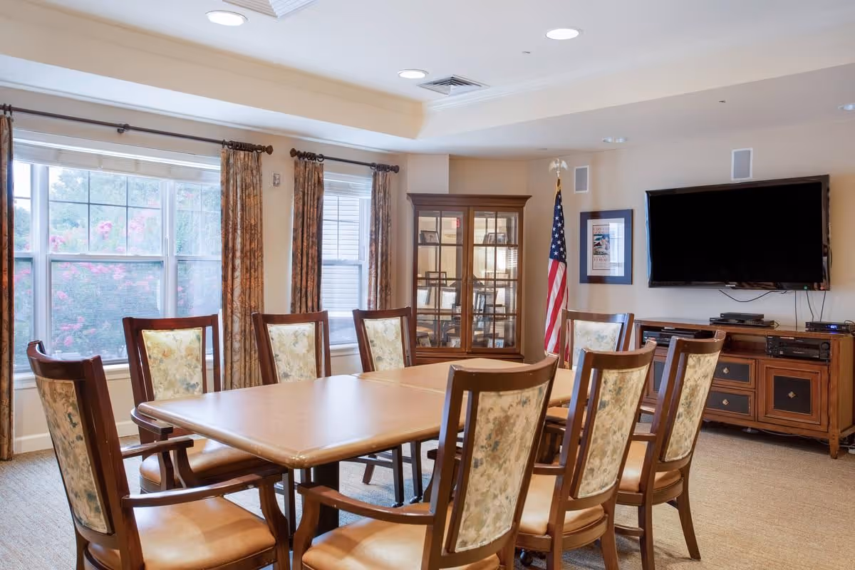 A well-lit meeting or dining room with a rectangular wooden table surrounded by eight upholstered chairs. The room features large windows with floral curtains, a glass display cabinet, an American flag, a wall-mounted flat-screen TV, and a wooden media console beneath the TV.