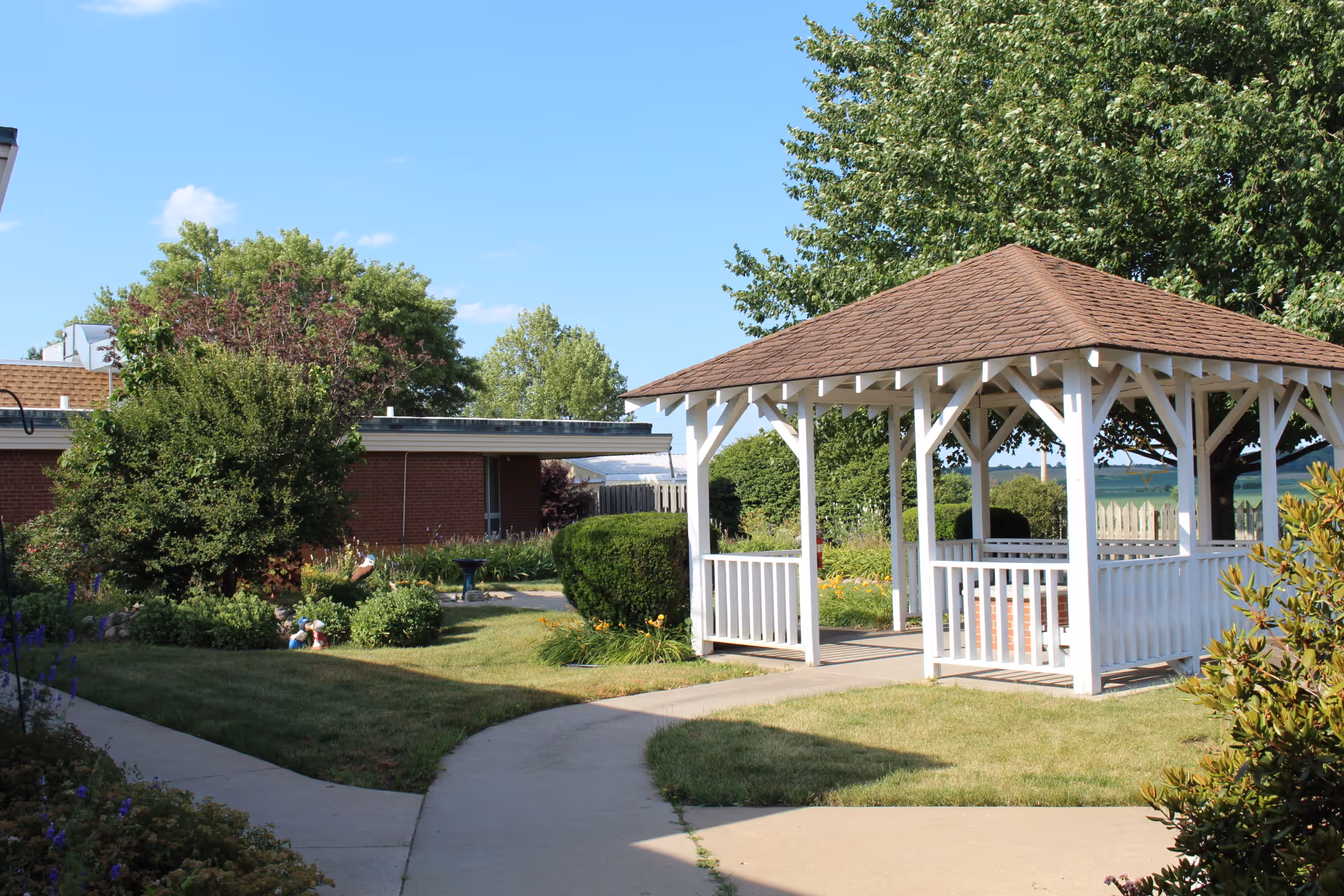 A sunny outdoor garden area at Sunny Acres Nursing Home featuring a white wooden gazebo with a brown shingled roof, surrounded by green grass, bushes, trees, and a paved walkway.
