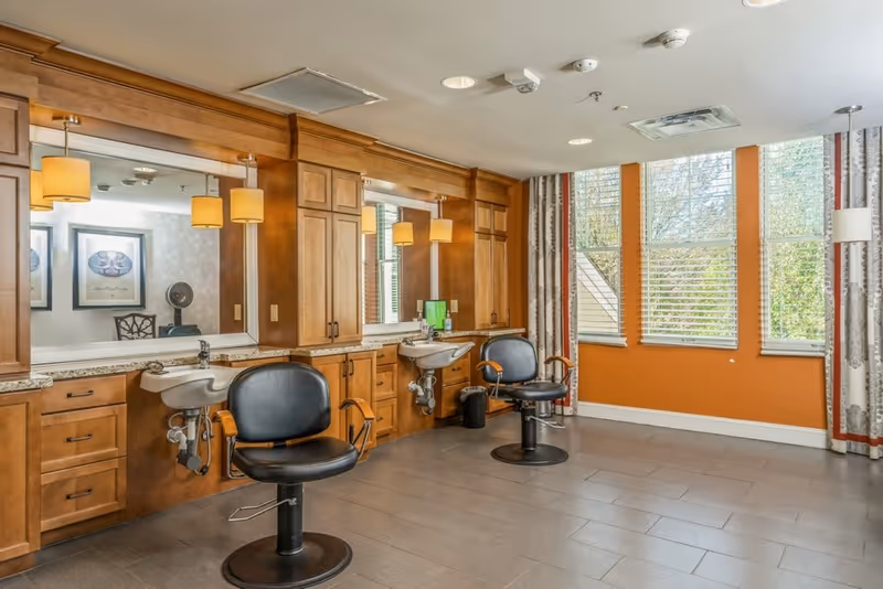 Interior view of a salon area in a senior living facility with two black salon chairs in front of two sinks and large mirrors mounted on wooden cabinetry. The room has large windows with blinds and patterned curtains, and warm lighting fixtures above the mirrors.
