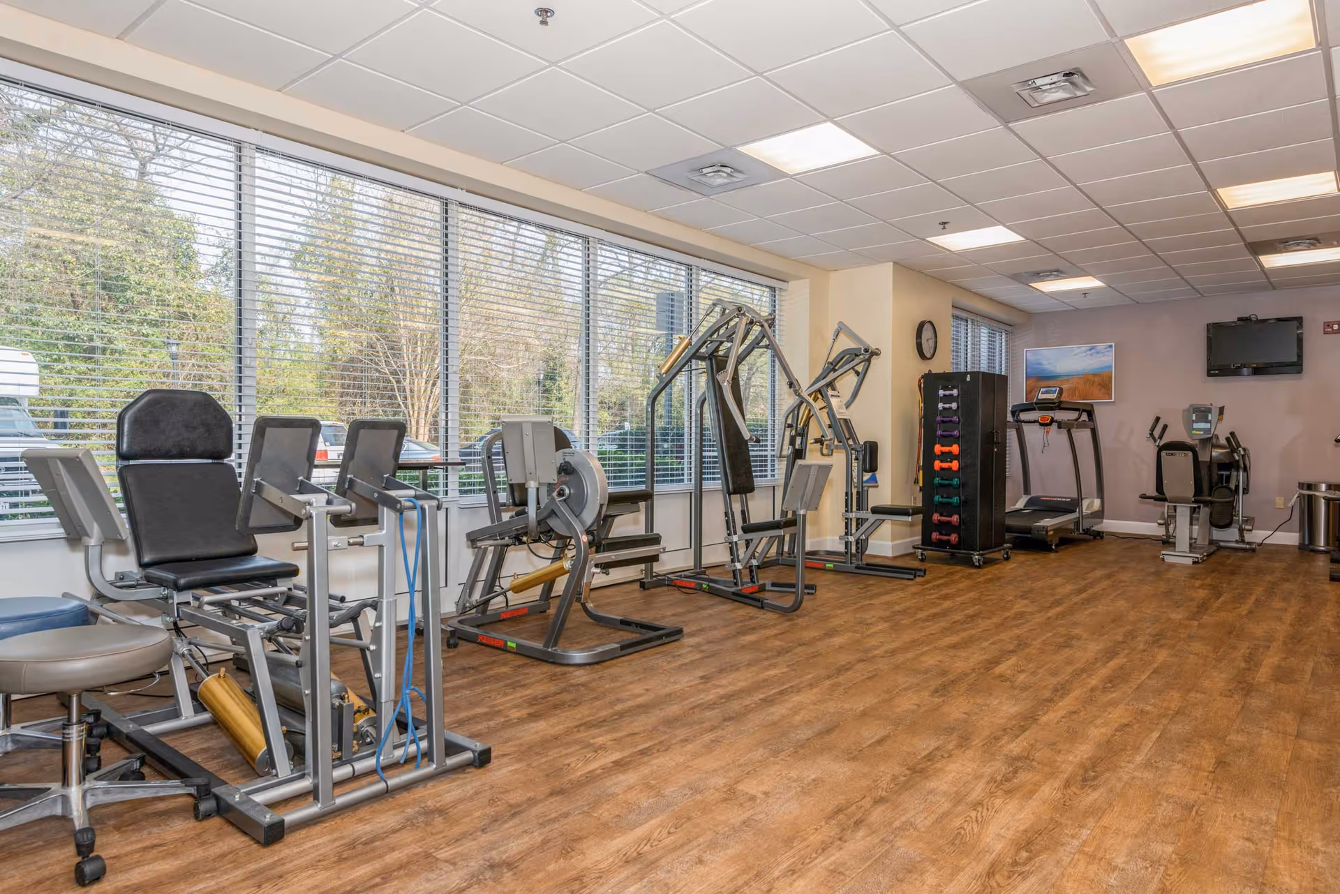A fitness room with various exercise equipment including leg press machines, a treadmill, a stationary bike, and a rack of colorful dumbbells. The room has large windows with blinds, wood-style flooring, and a wall clock.