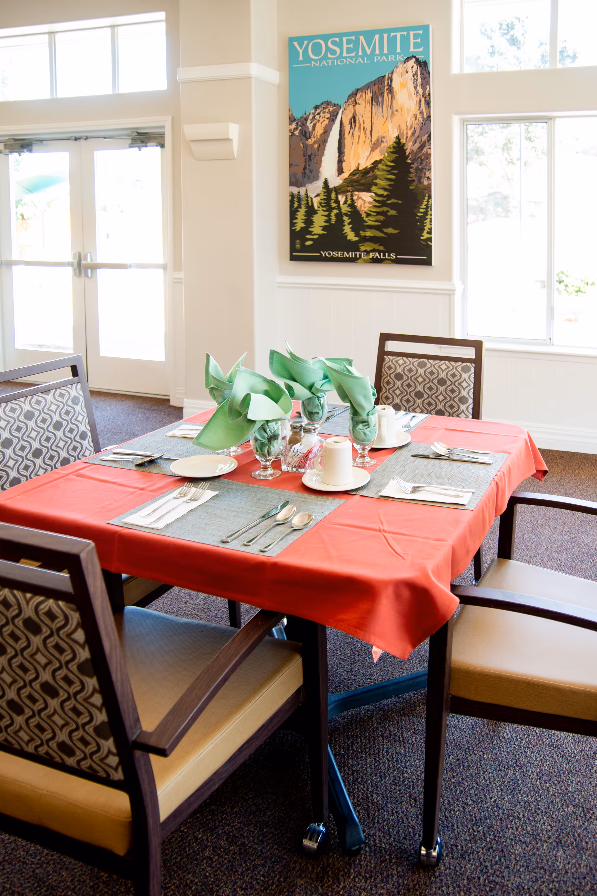 A dining table set for four with a red tablecloth and green folded napkins in glasses. The table has plates, cups, and silverware arranged neatly. Four chairs with patterned backs surround the table. A Yosemite National Park poster hangs on the wall, and there are large windows and a glass door letting in natural light.