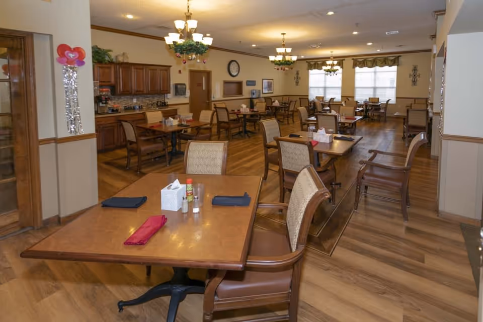 A spacious dining room in a senior living facility with multiple wooden tables and cushioned chairs arranged neatly. Each table has napkins, tissue boxes, and condiments. The room features wood flooring, beige walls, and large windows with valances allowing natural light. Ceiling lights with decorative greenery hang from the ceiling. A wooden cabinet and a clock are visible on the far wall.