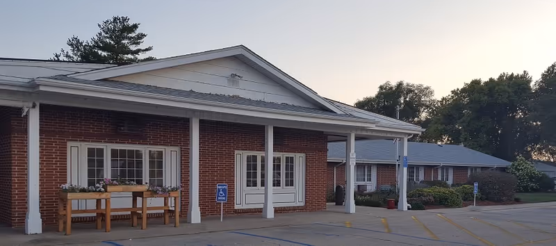 Exterior view of a single-story brick building with white trim and a covered entrance supported by white columns. There are flower boxes with purple flowers on a wooden stand near the windows. The parking lot in front has marked handicap parking spaces. Trees and shrubbery are visible in the background under a clear sky at dusk.