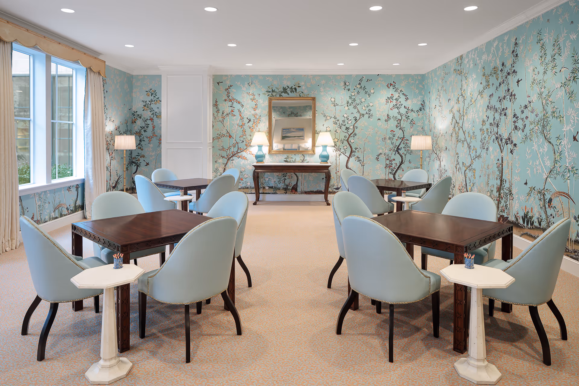 A bright and elegant room with light blue floral wallpaper featuring birds and branches. The room contains several dark wood square tables, each surrounded by four light blue upholstered chairs. There are two small white side tables with pencil holders. The room is well-lit with recessed ceiling lights and floor lamps. A large window with beige curtains allows natural light to enter. At the far end of the room, there is a wooden console table with two blue lamps and a framed mirror above it.