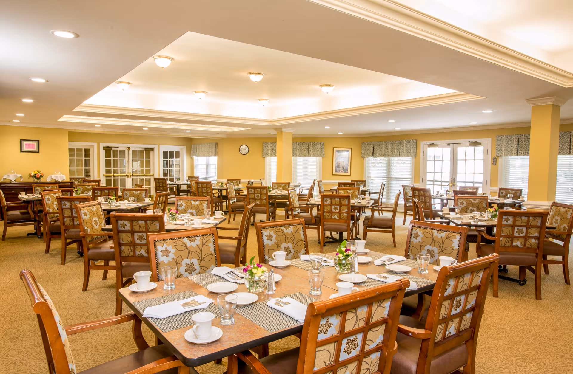 A spacious dining room with multiple wooden tables and chairs arranged neatly. Each table is set with white cups, plates, glasses, napkins, and small floral centerpieces. The room has warm yellow walls, large windows with patterned valances, and bright ceiling lights.