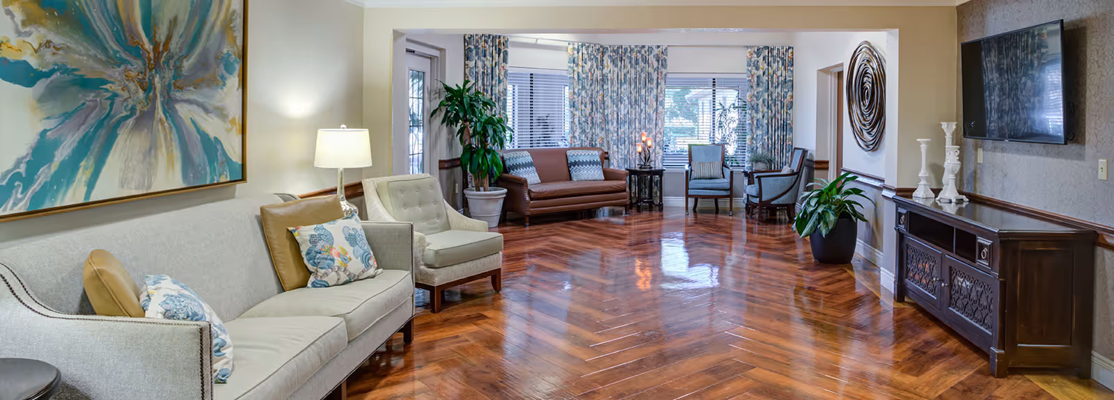 Spacious, well-lit common room with sofas and armchairs, decorative artwork, a wall-mounted TV, plants and polished herringbone hardwood floors.