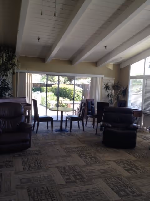 A cozy living room area with several chairs and a small round table near large windows that look out onto a garden. The room has a high ceiling with exposed beams and carpeted floor with a patterned design. There are plants near the windows and natural light fills the space.