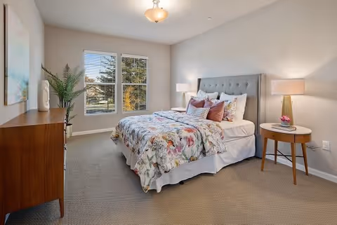 A bright and cozy bedroom with a large bed featuring a tufted gray headboard, floral bedding, and multiple pillows. There are two bedside tables with lamps on either side of the bed. A wooden dresser with a potted plant and decorative items is positioned against the left wall. Two large windows allow natural light to fill the room, and a piece of abstract art hangs on the wall above the dresser.