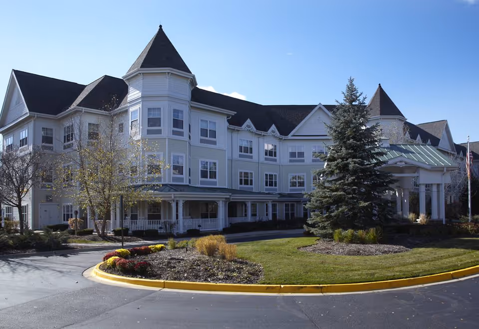 Exterior view of a large, multi-story senior living facility with white siding and dark roofing. The building features a turret and a covered entrance with columns. There is a landscaped roundabout with grass, bushes, and a large evergreen tree in front of the building under a clear blue sky.
