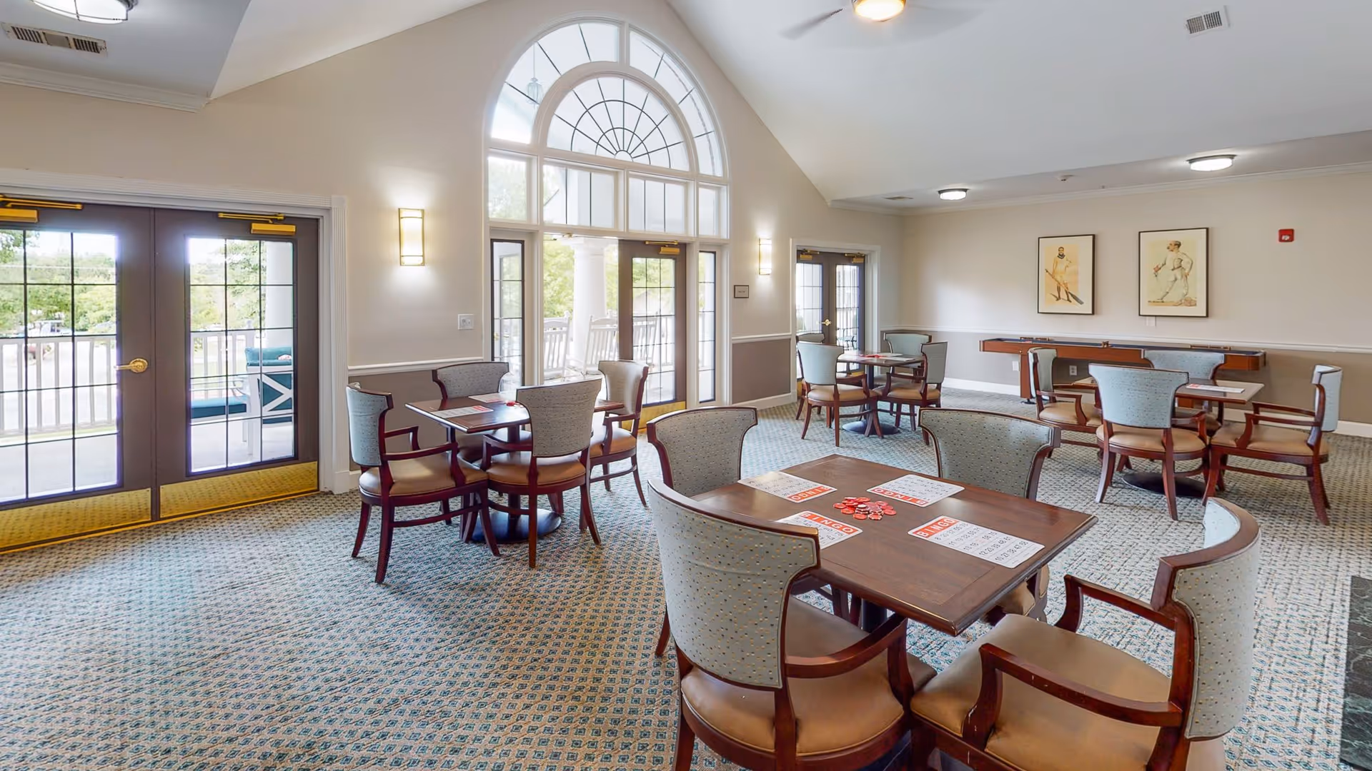 Bright communal dining and activity room with multiple tables and upholstered chairs, large arched windows and doors to an outdoor balcony.