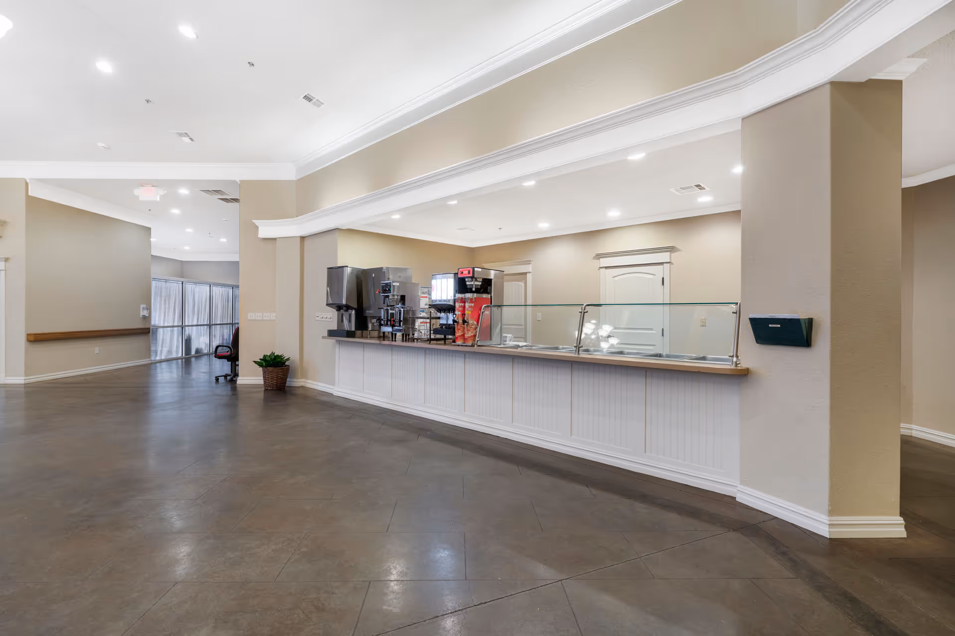 Interior view of a senior living facility lobby with a long food service counter, coffee machines, and an open seating area.