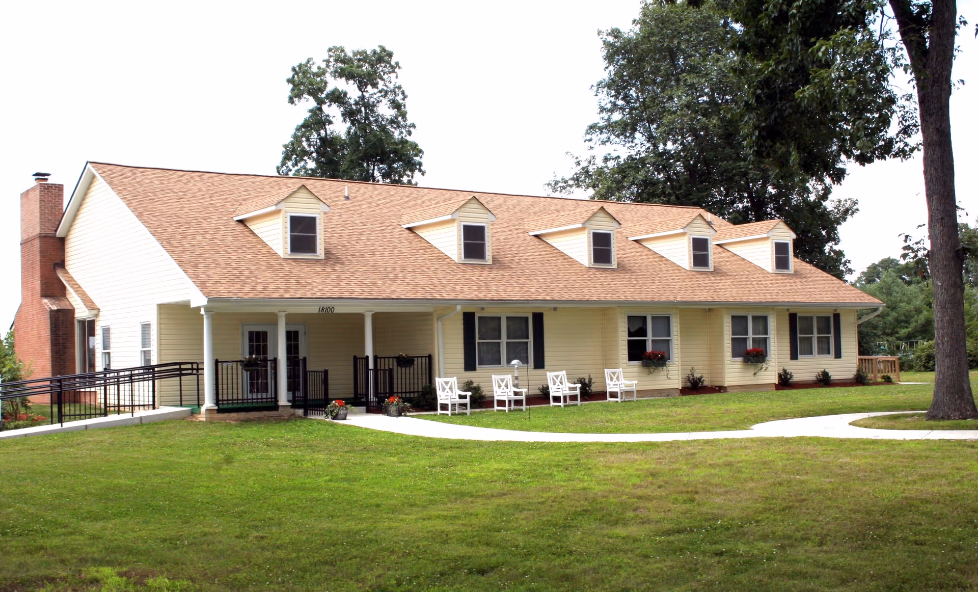 A single-story yellow building with a brown shingled roof and several dormer windows. The building has a covered porch with white columns and white chairs arranged outside. There is a paved walkway leading to the entrance and a grassy lawn in front with a large tree on the right side.