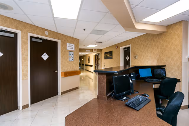 Reception desk area with computer monitors and chairs in a nursing and rehabilitation center hallway. The hallway has beige walls, tiled floors, and several closed doors with signs. There is a handrail along the wall and a ceiling with recessed lighting.