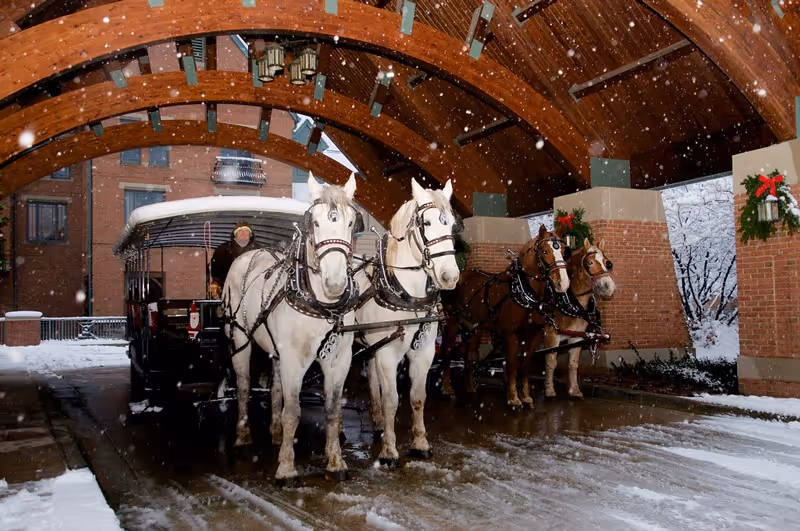 A winter scene showing a horse-drawn carriage with four horses standing under a wooden arched canopy at the entrance of a building. Snow is falling and accumulating on the ground and the roof. The building is decorated with holiday wreaths with red bows.