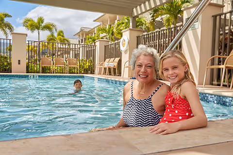 An elderly woman and a young girl smiling and sitting at the edge of a swimming pool with a boy swimming in the background. The pool area is surrounded by a fence, palm trees, and outdoor furniture under a clear sky.