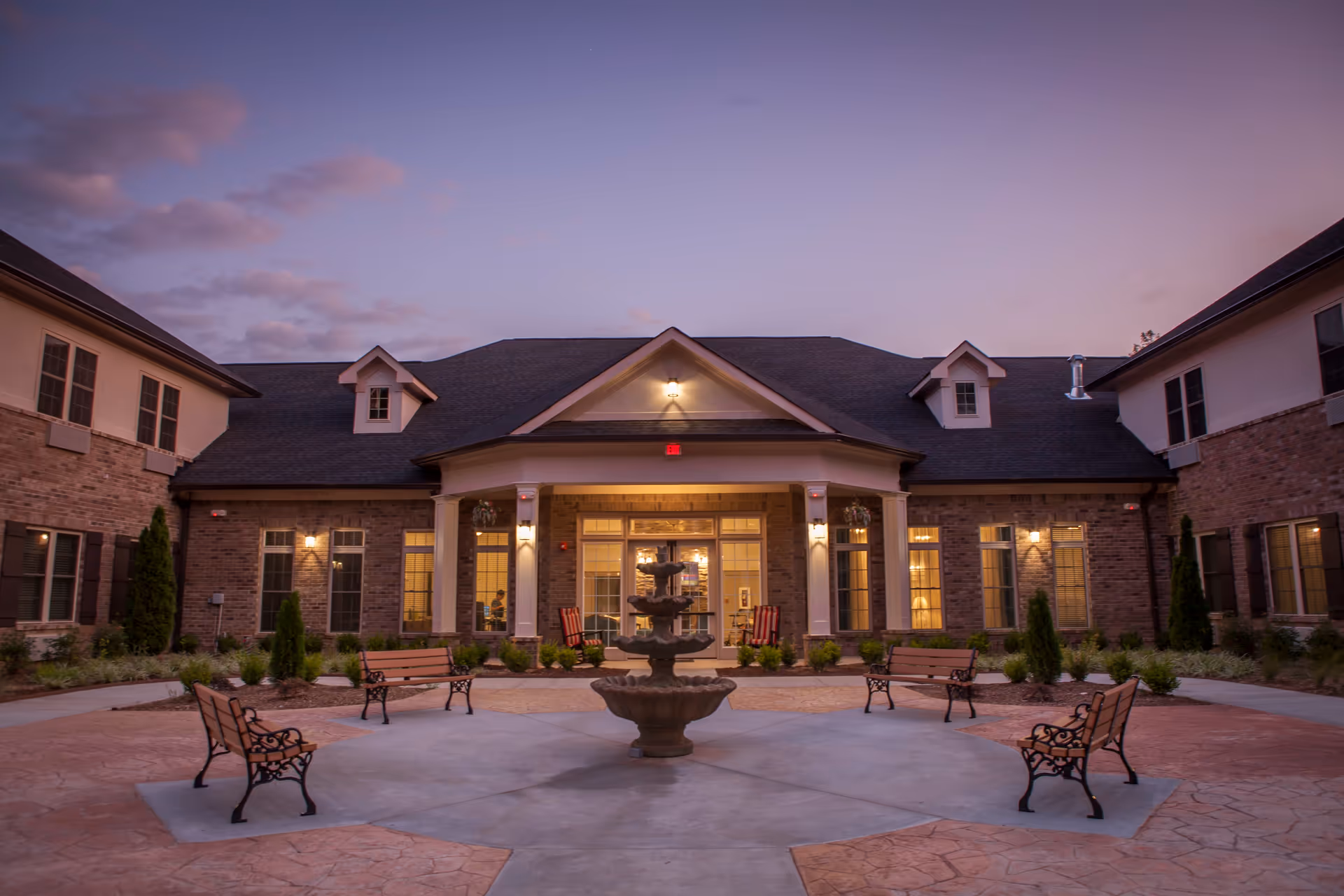 Courtyard with benches and a central fountain in front of the illuminated entrance of a senior living building at dusk.