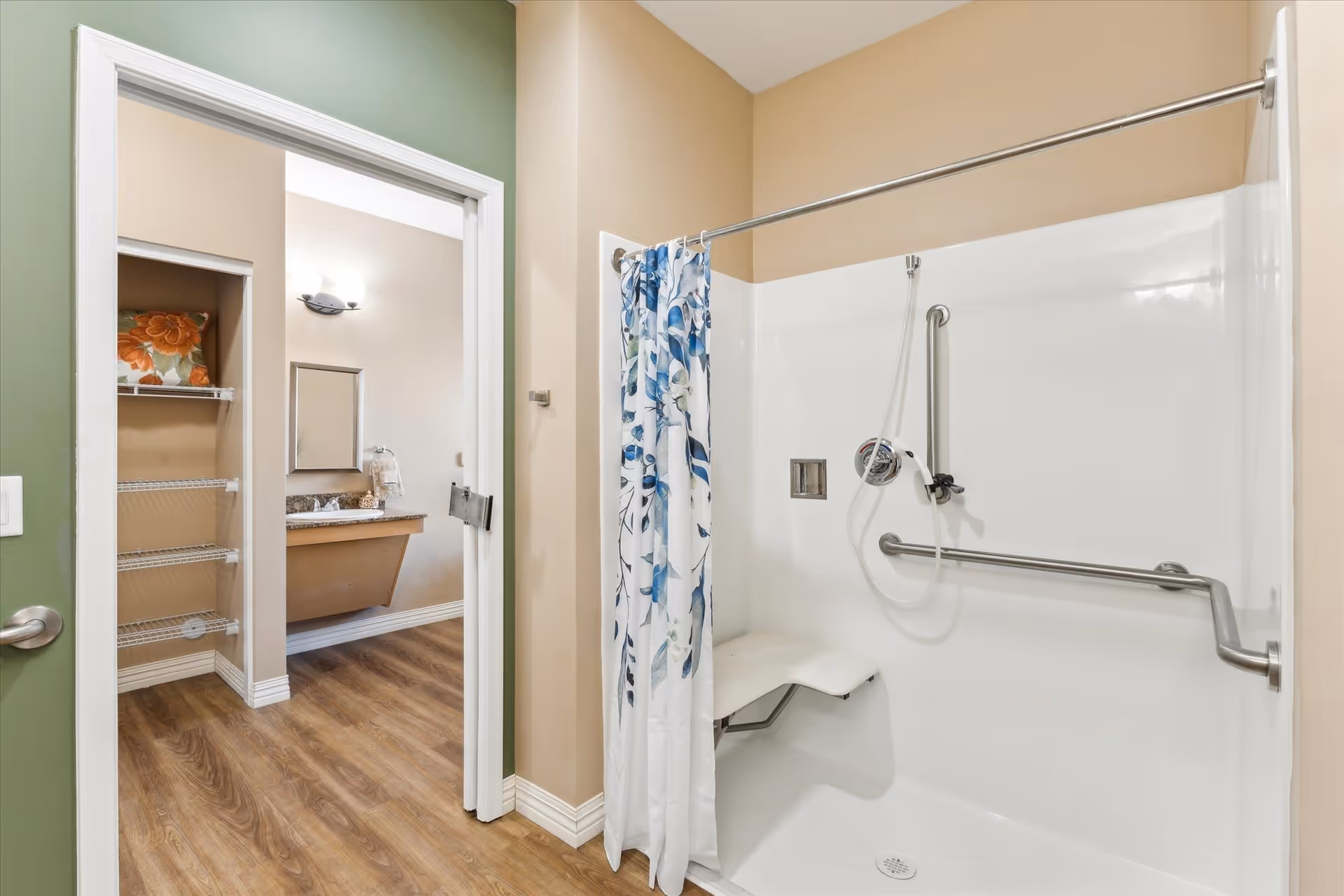A bathroom with a walk-in shower featuring a white fold-down seat, grab bars, and a floral shower curtain. The shower has a handheld showerhead. Adjacent to the shower is an open doorway leading to a vanity area with a mirror, countertop, and wall-mounted light fixture. The floor is wood-style laminate, and there is a small closet with wire shelving visible in the vanity area.