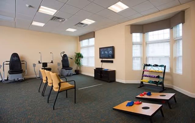 A well-lit exercise room with two seated exercise machines, three yellow chairs arranged in a row, a TV mounted on the wall above a small cabinet, a rack holding colorful dumbbells, and two cornhole boards with bean bags on the carpeted floor near large windows.